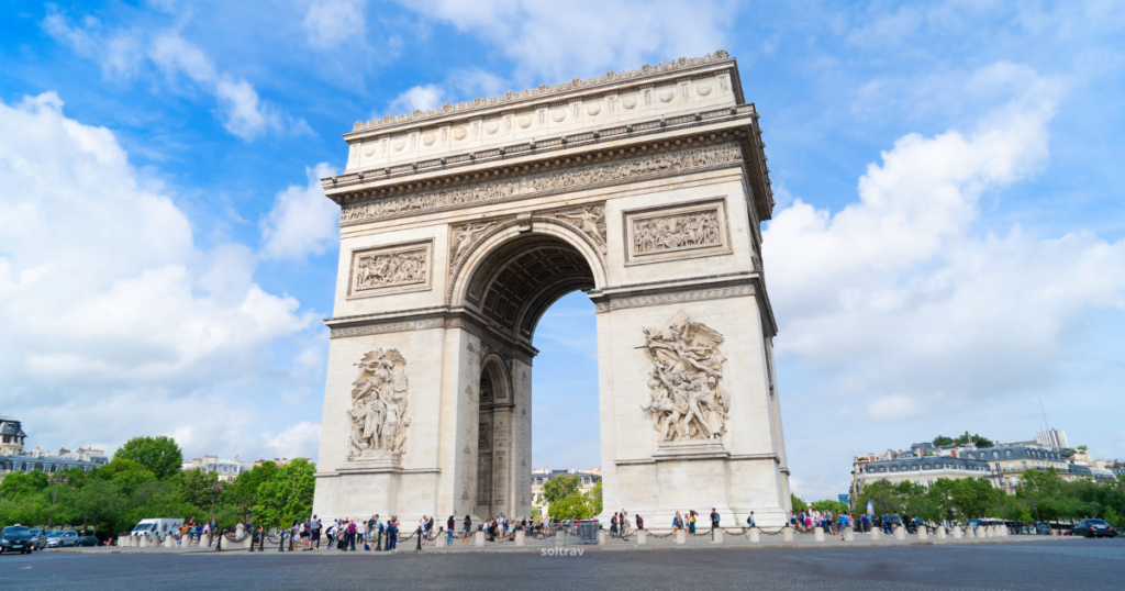 A majestic view of the Arc de Triomphe in Paris. The monument features intricate sculptures and relief on its façade, surrounded by a lively crowd of visitors. The sky is partly cloudy, adding to the vibrant atmosphere of this iconic landmark.