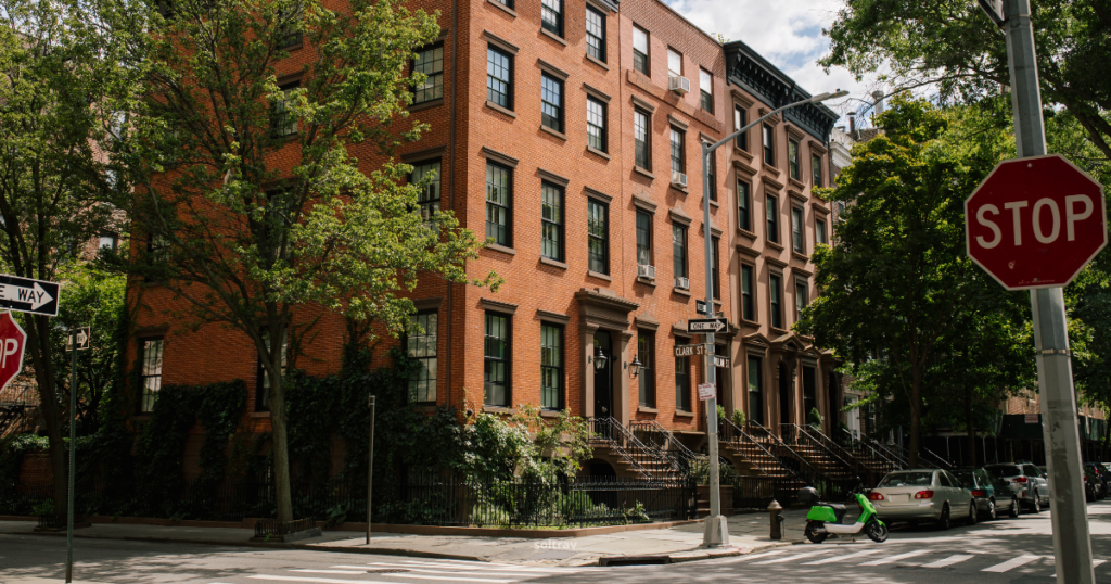 A charming street scene in Brooklyn featuring red brick buildings with large windows and decorative staircases. Street signs indicate 'Clark St.' and a 'One Way' direction, while a 'Stop' sign is prominently displayed. Lush trees provide greenery along the sidewalk.