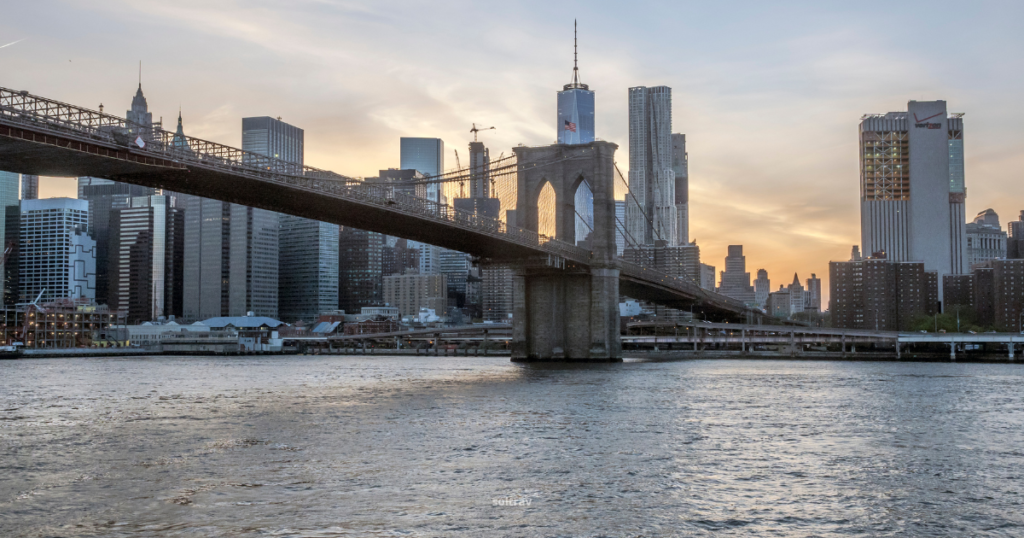 A view of the Brooklyn Bridge at sunset, stretching across the East River with the Manhattan skyline in the background. The bridge's intricate architecture contrasts with the modern skyscrapers, some of which are catching the warm glow of the setting sun. The water reflects the colors of the sky, creating a serene urban landscape.