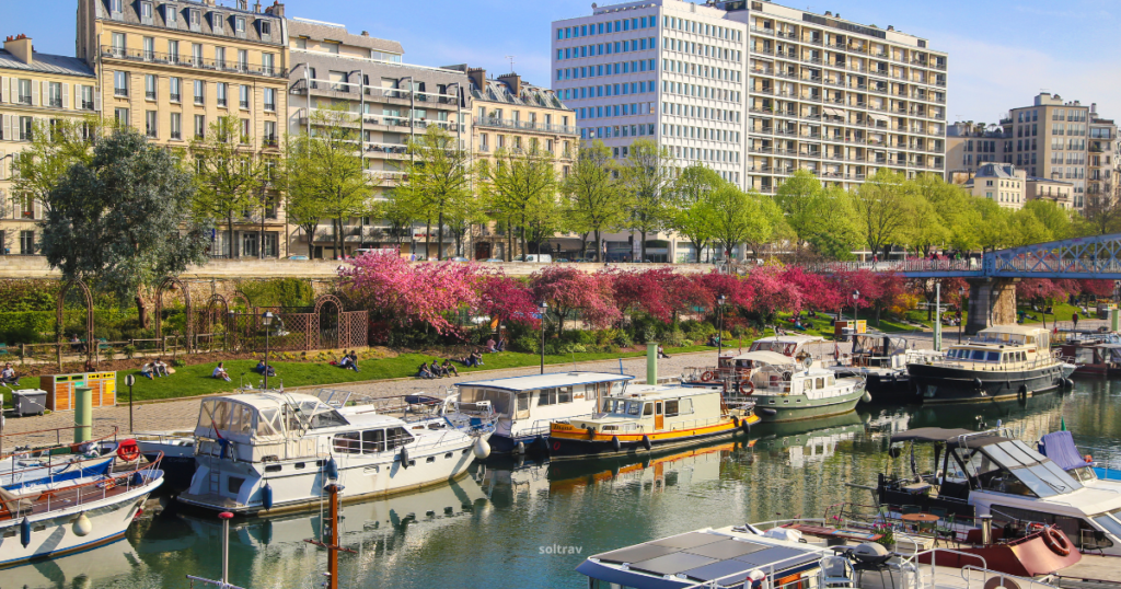 A vibrant view of Canal Saint-Martin in Paris, featuring a line of moored boats along the water's edge. Lush greenery and blooming pink flowers line the canal, with people relaxing on the grassy banks. Modern buildings rise in the background, creating a lively contrast with the serene waterway and its charming atmosphere.