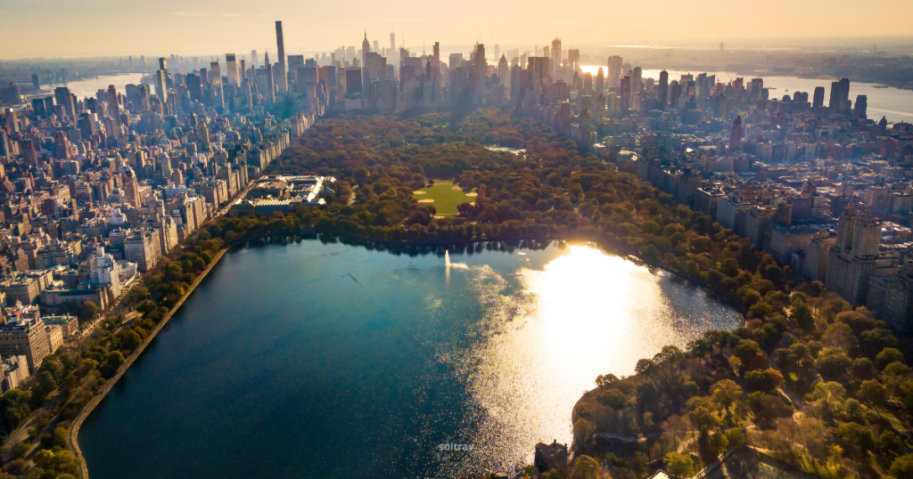 Aerial view of Central Park in New York City, surrounded by the city's skyline. The park features lush greenery and a large reflective body of water, with sunlight glistening on the surface. Skyscrapers loom in the background, showcasing the vibrant urban landscape. The scene captures the contrast between the natural space of the park and the bustling city beyond.