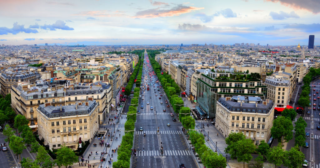 A panoramic view of the Champs-Élysées in Paris, showcasing the iconic boulevard lined with trees and bustling traffic. The wide avenue is flanked by elegant buildings, with a mix of shops and cafes visible. In the background, the skyline of Paris stretches out under a colorful sky, hinting at the city's vibrant atmosphere.