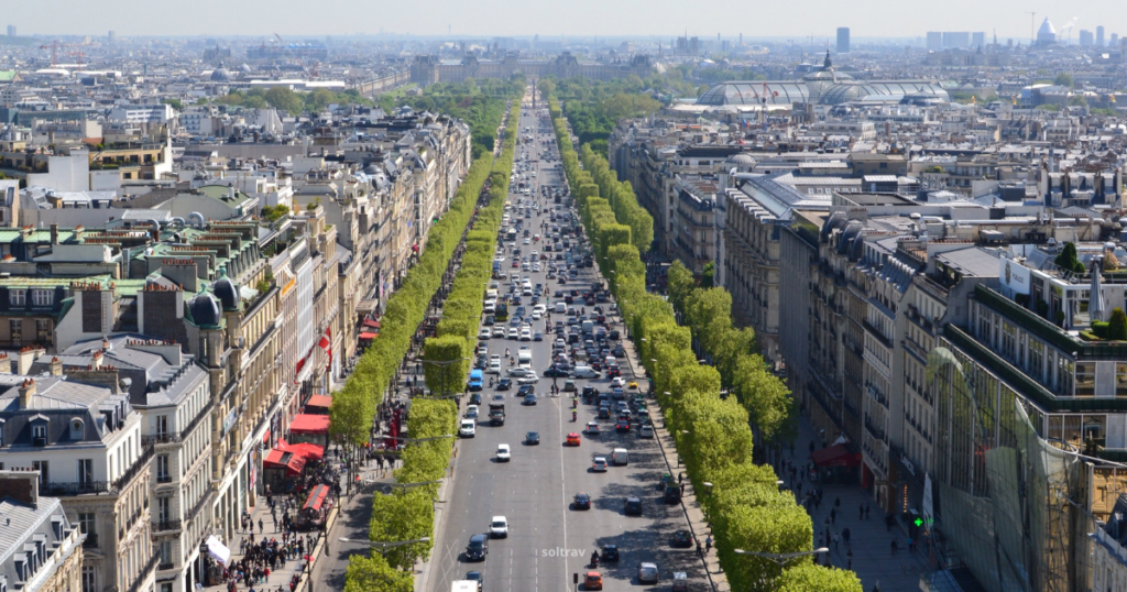 Aerial view of the Champs-Élysées in Paris, featuring a wide tree-lined avenue bustling with traffic and pedestrians. Elegant buildings with various architectural styles flank the street, along with shops and cafes. The scene is set against a clear blue sky, showcasing the vibrant atmosphere of this iconic location.