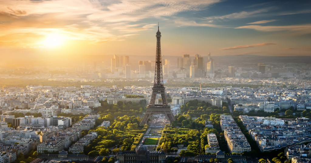 An aerial view of Paris during sunset, showcasing the Eiffel Tower standing prominently in the center. The surrounding cityscape is bathed in a warm, golden light, with rows of buildings, streets, and green parks. In the background, modern skyscrapers of the La Défense district are visible under a partly cloudy sky, adding contrast to the historical architecture in the foreground.