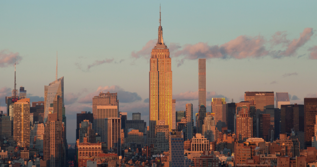 A close-up view of the Empire State Building at sunset, bathed in golden light. The iconic skyscraper stands prominently among the New York City skyline, surrounded by various modern buildings. The sky features soft clouds and a warm hue, enhancing the majestic presence of the Empire State Building as a symbol of the city.