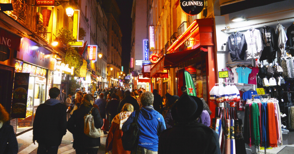 A bustling street in the Latin Quarter of Paris at night, filled with people walking along vibrant shops and restaurants. Colorful neon signs illuminate the scene, while outdoor dining areas create a lively atmosphere. The narrow street is lined with diverse eateries and shops, reflecting the area's energetic nightlife.