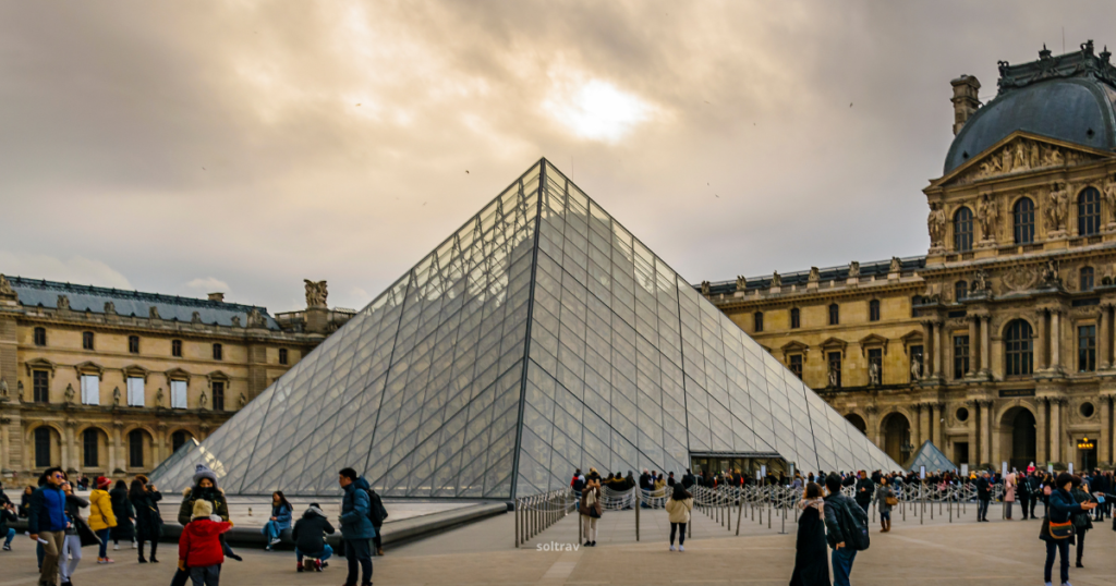 A view of the Louvre Pyramid in Paris, France, surrounded by visitors. The glass and metal pyramid stands prominently in the center of the courtyard, reflecting the cloudy sky above. The historic Louvre Palace buildings, with their ornate architectural details, form the backdrop, adding a contrast between modern and classical design elements.