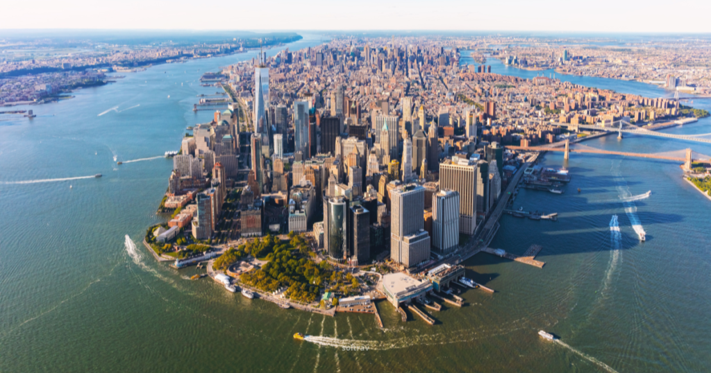 Aerial view of Manhattan, showcasing its iconic skyline with numerous skyscrapers and the waterfront. The surrounding waters are dotted with boats, and green spaces are visible along the shoreline, creating a vibrant urban landscape.