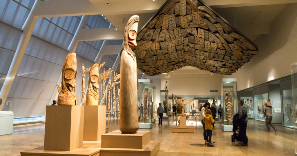 An interior view of an art museum featuring a collection of wooden sculptures and artifacts. Prominent among the exhibits are tall, intricately carved totem poles and masks displayed on pedestals. The museum's modern architecture is highlighted by large windows and a unique ceiling design. Visitors can be seen exploring the space, adding to the vibrant atmosphere of cultural appreciation.