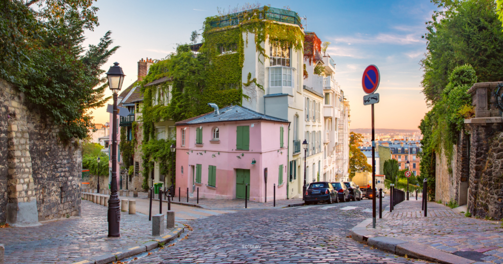 A charming street in Montmartre, Paris, featuring a cobblestone path winding past quaint buildings adorned with greenery. A small pink house with green shutters stands out among the surrounding structures. Street lamps and traffic signs add to the picturesque atmosphere, with a soft sunset glow illuminating the scene.