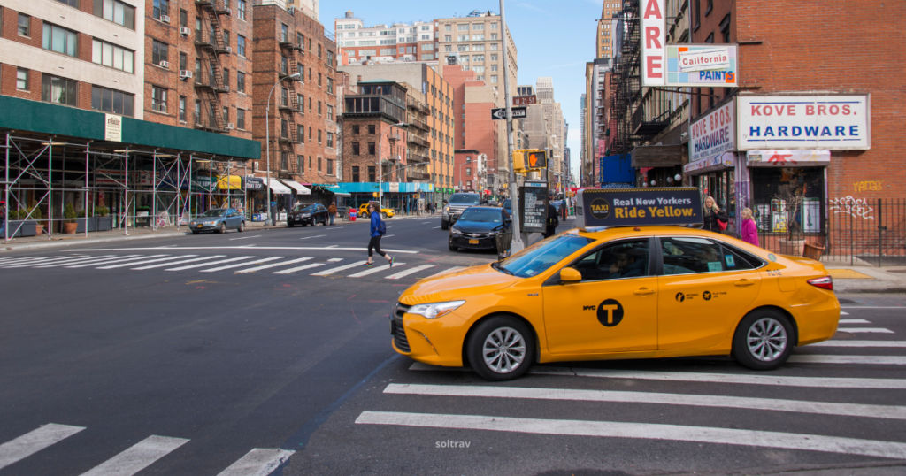 A yellow taxi making a turn at an intersection in New York City. The scene features brick buildings, street signs, and pedestrians crossing the street, with a sign on the taxi roof reading 'Real New Yorkers Ride Yellow.'