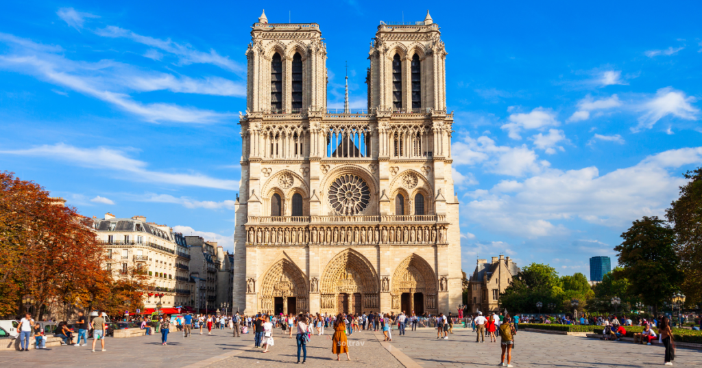 A bright day at Notre-Dame Cathedral in Paris, showcasing its iconic Gothic architecture. The façade features two tall towers and intricate sculptures. A crowd of visitors gathers in the foreground, surrounded by trees with autumn foliage and a clear blue sky.