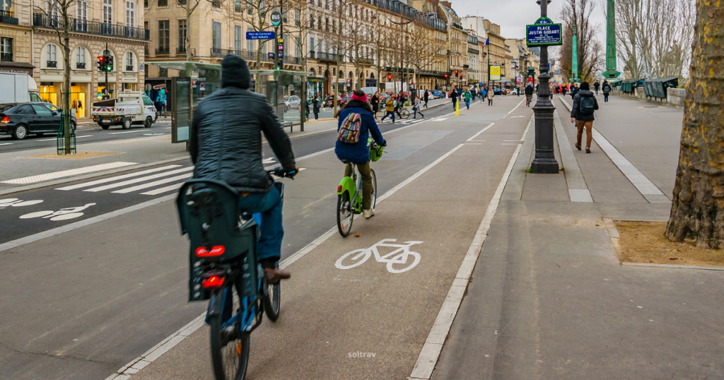 A busy bike lane in Paris, featuring cyclists riding alongside a bustling street. One cyclist is on a blue bike, while another rides a bright green bike. The sidewalk is lined with pedestrians and trees, and traffic moves in the background. Street signs and a bus stop add to the urban atmosphere of this vibrant city.