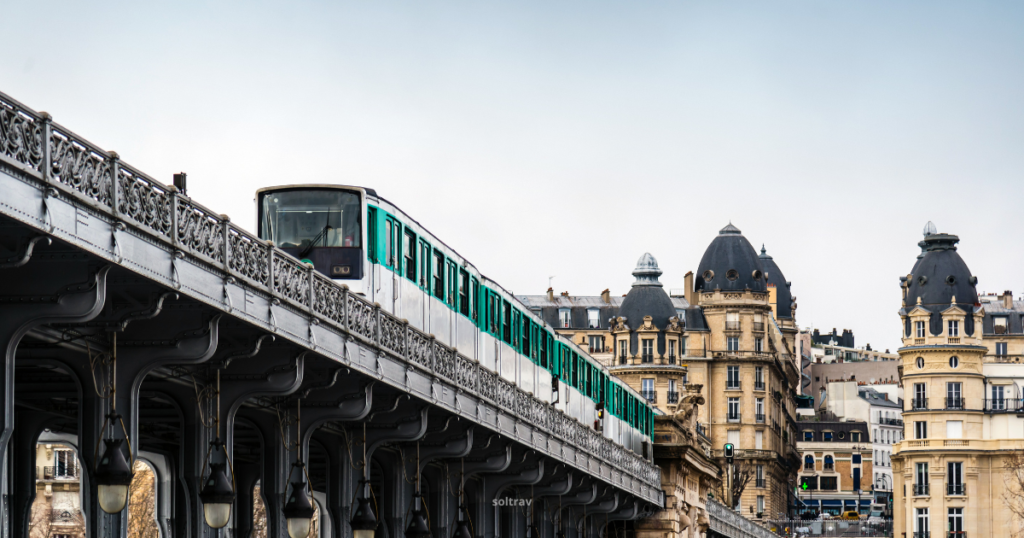 A Paris metro train travels along an elevated track, with ornate ironwork visible beneath. The train features a sleek design with green accents. In the background, traditional Parisian buildings with domed roofs create a charming urban landscape, set against a cloudy sky.