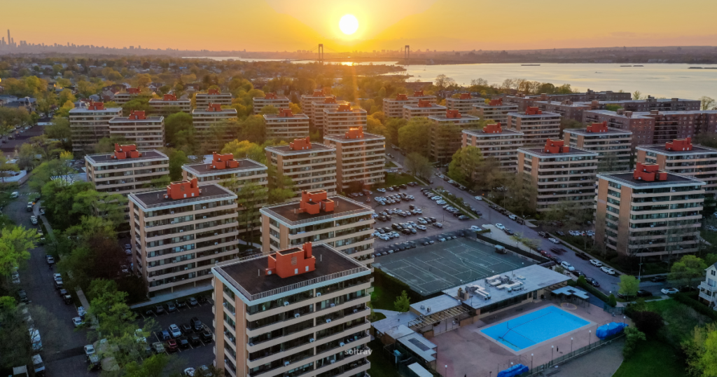 Aerial view of residential buildings in Queens, New York, during sunset. The scene includes multiple apartment complexes with red rooftops, a visible swimming pool, and tennis courts. The distant skyline and the water provide a picturesque backdrop as the sun sets on the horizon.