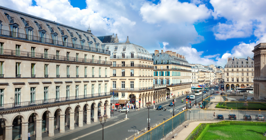 A view of Rue de Rivoli in Paris, highlighting elegant architecture with classic Parisian buildings lining the street. The facades feature arched windows and intricate detailing, while bustling traffic and pedestrians fill the road. The sky is bright with fluffy clouds, adding to the vibrant atmosphere of this iconic avenue.