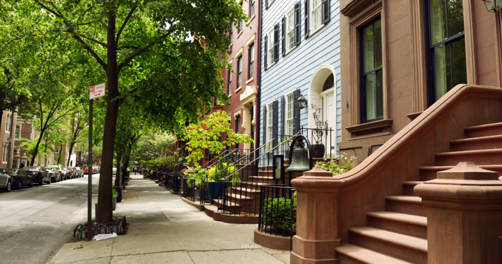 A peaceful New York City sidewalk lined with trees and brownstone buildings. The scene features steps leading to homes, a 'No Parking' sign, and a bell mounted on a post, creating a charming urban atmosphere.