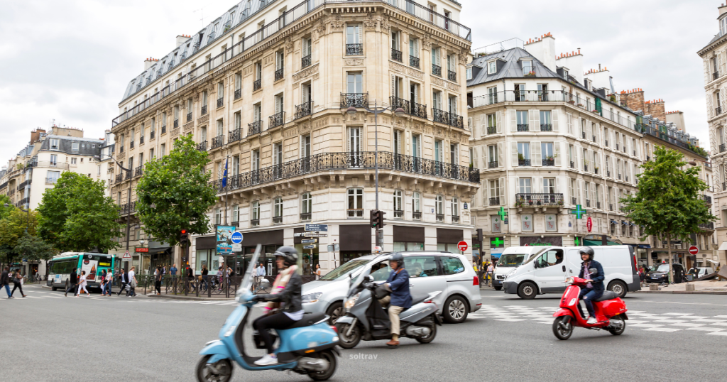 A lively street scene in St-Germain-des-Prés, Paris, showcasing a bustling intersection. Motorcycles and cars navigate the road, while pedestrians walk along the sidewalks lined with leafy trees and classic Parisian architecture. The buildings feature intricate balconies, and various signs indicate shops and amenities in this vibrant neighborhood.