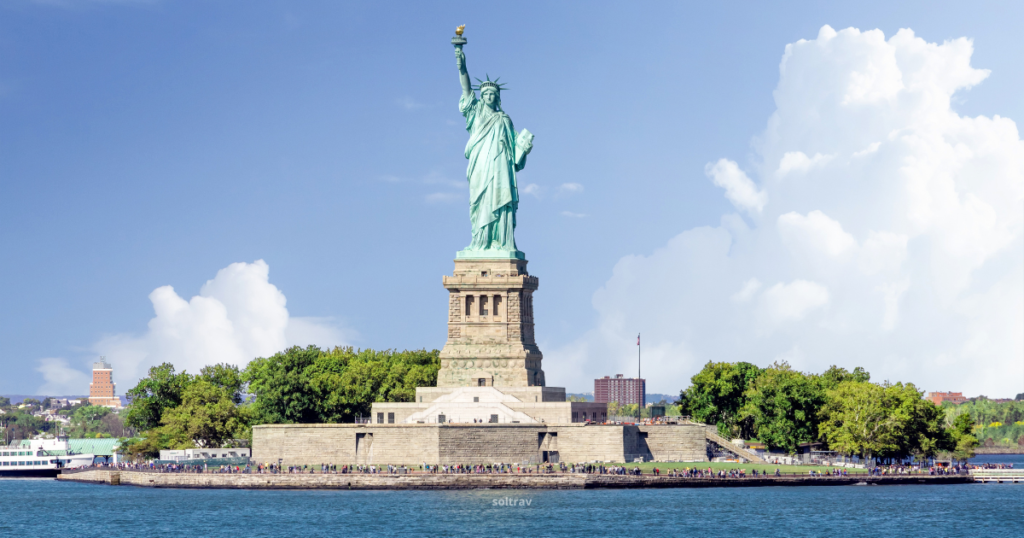 A view of the Statue of Liberty standing tall on Liberty Island, with her torch raised high. The statue is surrounded by lush greenery and a crowd of visitors at its base. In the background, a bright blue sky with fluffy white clouds enhances the scene, while the waters of the harbor gently reflect the statue and its pedestal. The image captures the iconic symbol of freedom and democracy.