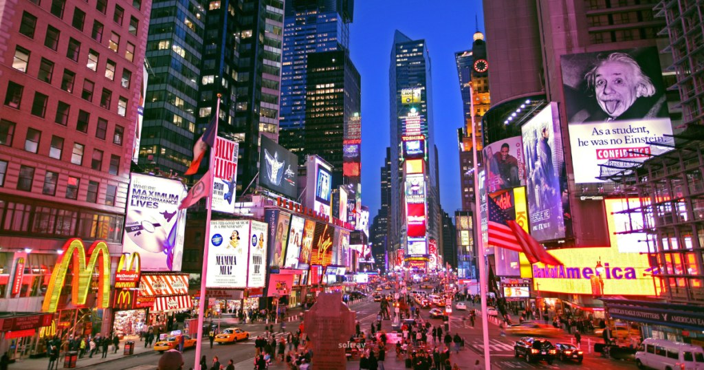 A bustling view of Times Square in New York City at dusk, showcasing bright, colorful advertisements and billboards. The street is filled with pedestrians and yellow taxis, with the iconic McDonald's sign visible in the foreground. Tall skyscrapers surround the area, and American flags are prominently displayed. The vibrant atmosphere captures the energy of the city.