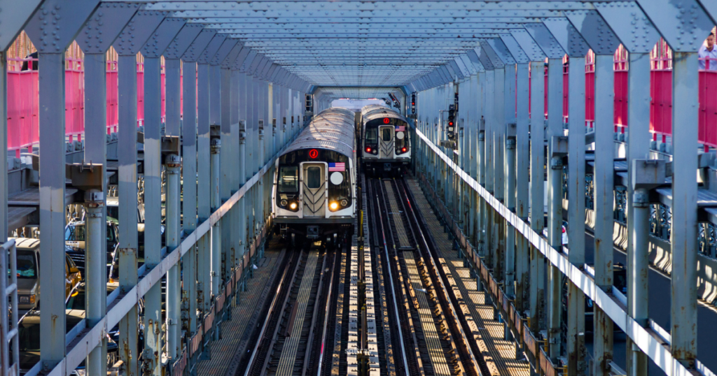 Two New York City subway trains approaching each other on elevated tracks, framed by the steel structure of the subway overpass. Bright sunlight casts shadows on the tracks, while colorful barriers line the sides of the platform.