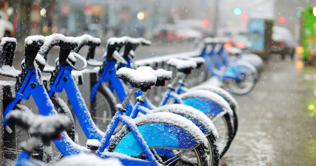 A row of blue rental bikes covered in a layer of fresh snow, set against a blurred city background. Snowflakes fall gently, creating a wintry scene on a bustling New York City street.