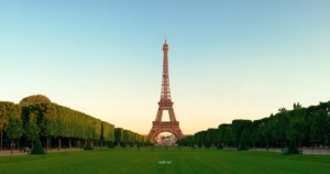 A view of the Eiffel Tower at sunset, framed by symmetrical rows of lush green trees and an expansive lawn in Paris, France