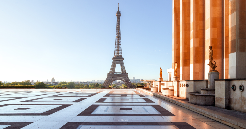 A view of the Eiffel Tower in Paris, France, from the Trocadéro plaza. The foreground features a large tiled terrace with geometric patterns. To the right, there are statues and the columns of a building, warmly lit by sunlight. The Eiffel Tower stands prominently in the center, with a clear blue sky above.