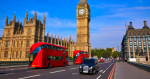 A vibrant view of the Houses of Parliament and Big Ben in London on a sunny day. Two modern red double-decker buses are crossing Westminster Bridge, along with a classic black London taxi. The background features the iconic architecture of Parliament, and the Union Jack is flying on a nearby building under a bright blue sky.