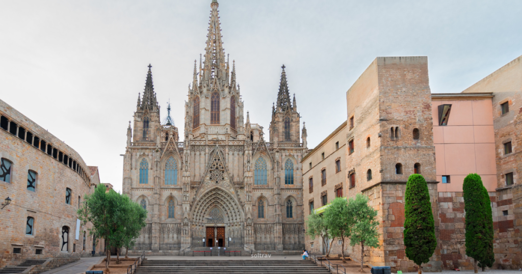 A picturesque view of the Cathedral of Barcelona, set in the Gothic Quarter. The cathedral features an elaborate Gothic facade with tall spires and intricate stone carvings. In the foreground, a wide staircase leads to the entrance, framed by neatly trimmed trees. To the left, a historic stone building adds to the medieval atmosphere, while soft clouds linger in the sky.