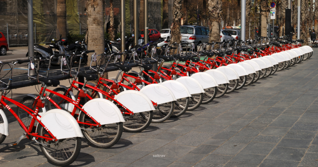 A row of red rental bicycles lined up on a paved sidewalk in Barcelona. Each bicycle has a distinctive white fender and is positioned neatly along the curb, with several parked cars visible in the background. The scene captures the vibrant urban atmosphere, emphasizing the accessibility of cycling in the city.