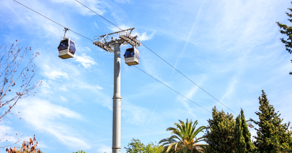 A Montjuïc cable car gliding along its overhead cables against a bright blue sky. The cable car features a sleek design with glass panels, and two cabins are visible suspended in the air. Below, lush greenery and palm trees are framed by the clear sky, creating a vibrant and scenic atmosphere.