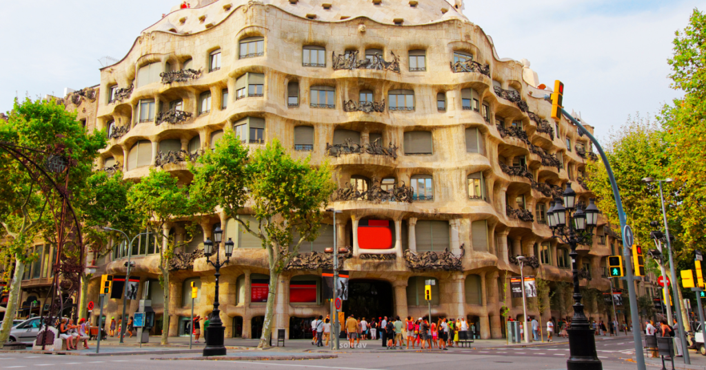 A street view of Casa Milà, also known as La Pedrera, in Barcelona. The building features an undulating stone facade with decorative wrought-iron balconies and windows. In the foreground, a crowd of people is gathered, and traffic lights and street lamps line the street. Lush trees provide greenery, contrasting with the unique architectural design.