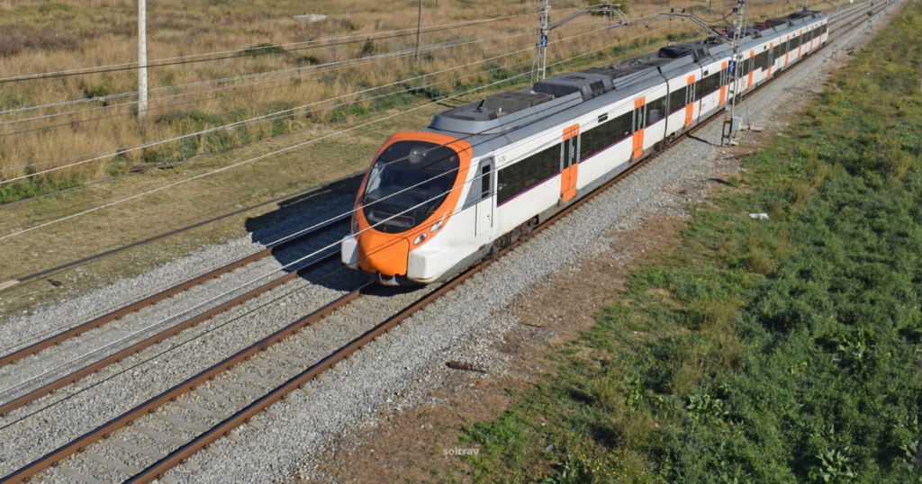 A passenger train traveling through the countryside near Barcelona, featuring a modern design with a white body and an orange front. The train is positioned on dual tracks surrounded by lush greenery and tall grass, with power lines running alongside. The scene captures the blend of urban transport and natural landscape.