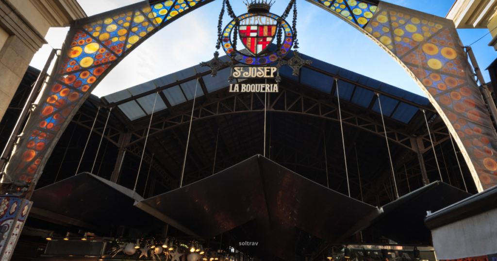 A striking view of the entrance to La Boqueria Market, featuring a colorful stained glass canopy adorned with vibrant designs. The sign above reads 'Mercat de Sant Josep de la Boqueria,' welcoming visitors to this famous market. The structure showcases a blend of modern and traditional architectural elements, hinting at the bustling atmosphere and rich offerings inside.