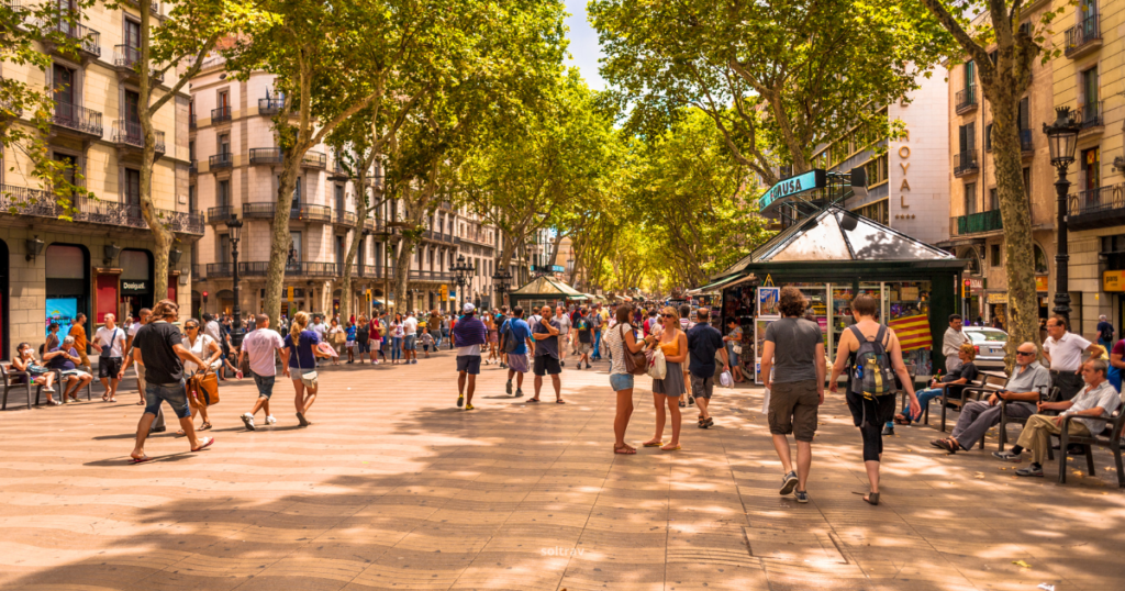 A bustling scene on La Rambla, a popular tree-lined street in Barcelona. The walkway is filled with a diverse crowd of people strolling, chatting, and enjoying the vibrant atmosphere. On either side, shops and cafes line the street, with outdoor seating inviting visitors to relax. Lush green trees provide shade, enhancing the lively ambiance of this iconic location.