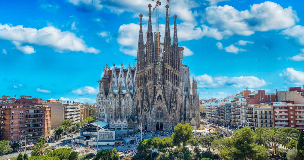 A panoramic view of the Sagrada Família, a famous basilica in Barcelona, Spain. The intricate architecture features tall spires and detailed stonework, set against a bright blue sky with fluffy white clouds. In the foreground, trees and people can be seen, while residential buildings line the street nearby.