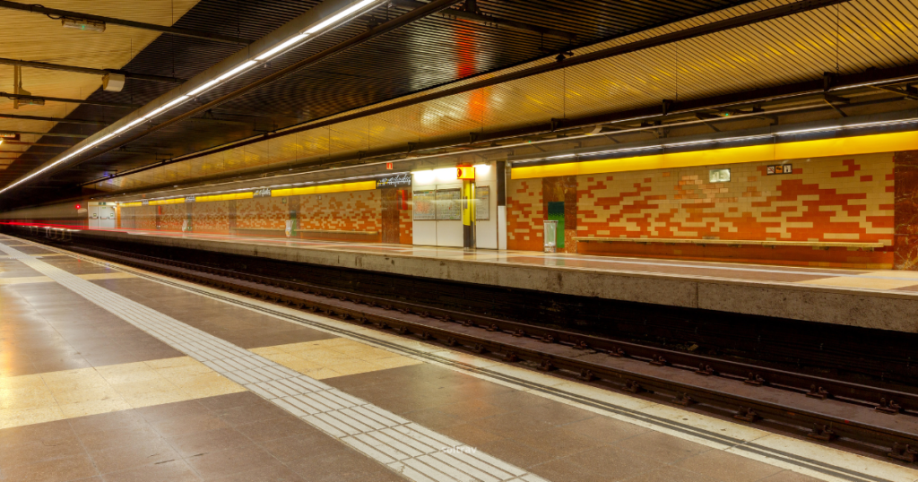 An empty platform inside a Barcelona Underground Metro Station, featuring modern architectural elements. The walls are adorned with a patterned tile design in warm colors, while the ceiling is lined with sleek metal panels. Bright lighting illuminates the space, highlighting the clean and spacious area, with tracks visible in the background.