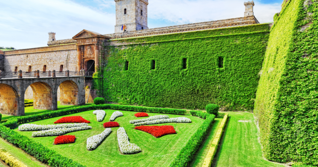 A vibrant view of the gardens at Montjuïc, featuring a beautifully designed flower bed in the shape of a sunburst, made up of red and white flowers. The lush greenery surrounds the garden, complemented by the imposing stone walls of the Montjuïc castle in the background. The scene is brightened by a clear blue sky, creating a serene and picturesque atmosphere.