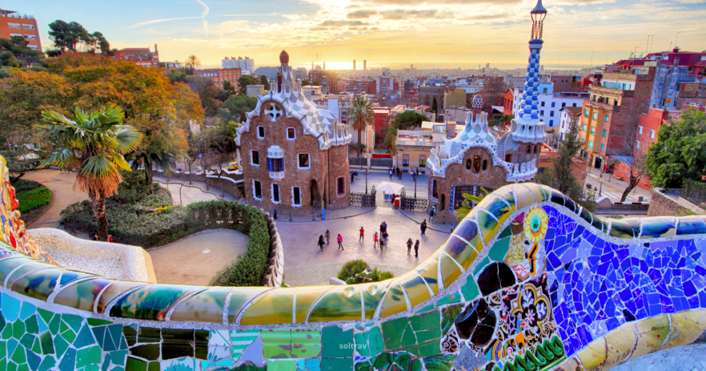 A vibrant view from Park Güell in Barcelona, showcasing colorful mosaic tiles in the foreground. The scene features unique architectural structures, including whimsical buildings with organic shapes. In the background, a sunset casts a warm glow over the city, with trees and people walking along the pathways, highlighting the park's artistic design.