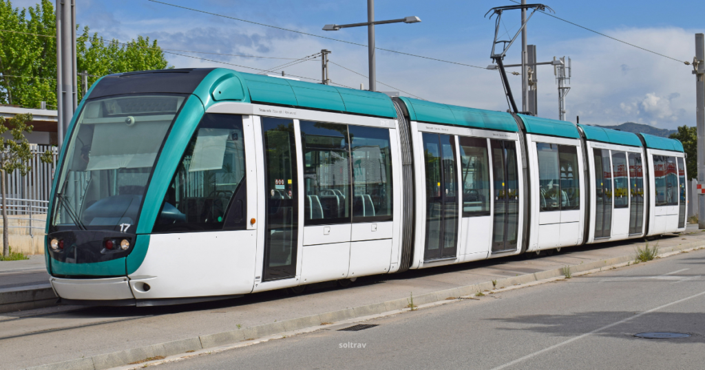 A modern Barcelona tram parked at a stop, showcasing its sleek design with a predominantly white body and teal accents. The tram features large windows and a streamlined shape, set against a backdrop of clear blue skies and green trees. The urban environment highlights the tram's role in public transportation.