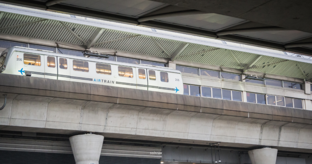 View of the AirTrain at JFK Airport, elevated above the ground on a track. The train, marked with the AirTrain logo, is in motion along its route. The background features the modern architecture of the airport terminal, with a concrete structure supporting the tracks, emphasizing the efficient transportation system connecting travelers to various airport locations.