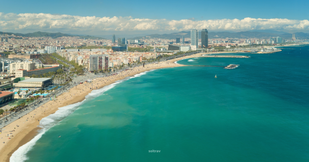 A panoramic view of Barceloneta Beach in Barcelona, showcasing a wide sandy shoreline bordered by gentle waves. Sunbathers and beachgoers can be seen enjoying the sun, while colorful umbrellas dot the landscape. In the distance, the city skyline rises with a mix of modern and historic architecture. The scene captures a lively summer atmosphere, reflecting the vibrant beach culture of Barcelona.