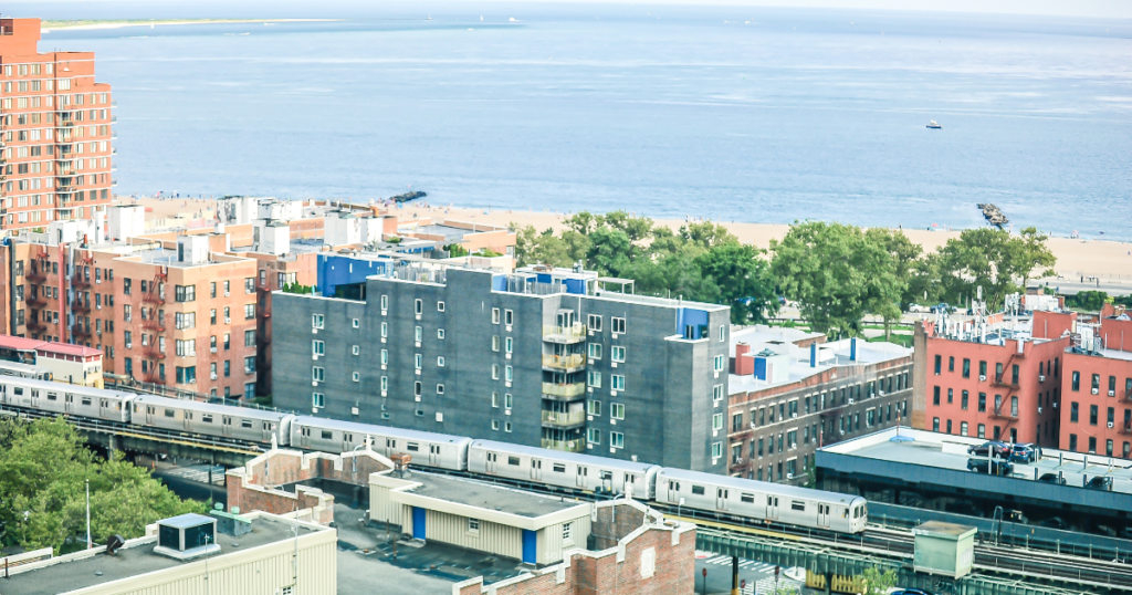 View of a Brooklyn neighborhood overlooking the beach and ocean. The image features a mix of residential buildings in various colors, with a subway train passing through the area. Lush green trees are interspersed among the buildings, and the sandy beach and calm waters are visible in the distance, creating a vibrant urban coastal scene.