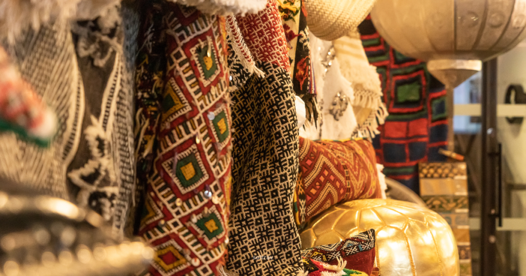 Close-up view of a textile shop in Chelsea Market, New York City, showcasing a variety of colorful fabrics and textiles. Intricate patterns and designs are displayed on hanging garments and textiles, with decorative items like golden cushions and lanterns visible. The vibrant colors and textures create a rich and inviting atmosphere, highlighting the shop's diverse offerings.