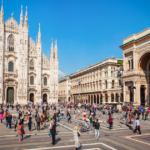 A wide-angle view of the Duomo di Milano, the stunning Gothic cathedral located in the heart of Milan. The intricate façade features numerous spires and statues, showcasing remarkable architectural detail. In the foreground, a bustling plaza is filled with people, some walking and others sitting, enjoying the vibrant atmosphere. Nearby, elegant buildings and a historic arch add to the charm of the scene, with clear blue skies overhead.