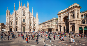 A wide-angle view of the Duomo di Milano, the stunning Gothic cathedral located in the heart of Milan. The intricate façade features numerous spires and statues, showcasing remarkable architectural detail. In the foreground, a bustling plaza is filled with people, some walking and others sitting, enjoying the vibrant atmosphere. Nearby, elegant buildings and a historic arch add to the charm of the scene, with clear blue skies overhead.