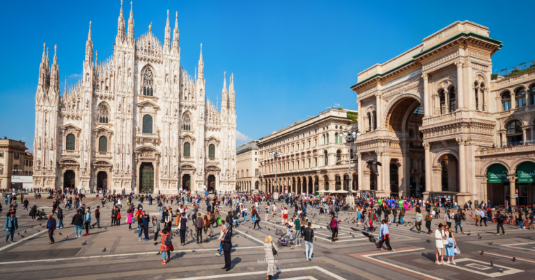 A wide-angle view of the Duomo di Milano, the stunning Gothic cathedral located in the heart of Milan. The intricate façade features numerous spires and statues, showcasing remarkable architectural detail. In the foreground, a bustling plaza is filled with people, some walking and others sitting, enjoying the vibrant atmosphere. Nearby, elegant buildings and a historic arch add to the charm of the scene, with clear blue skies overhead.