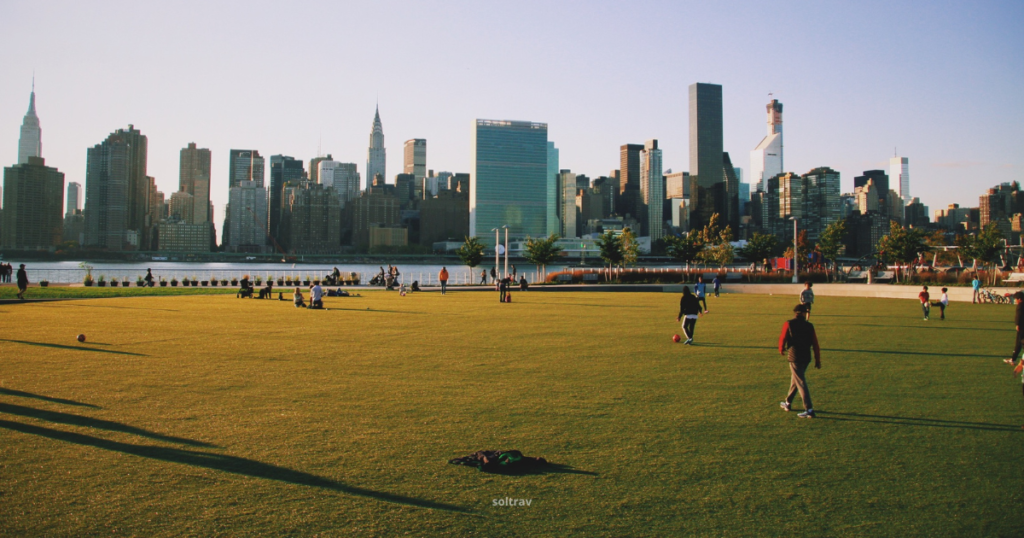 View of Gantry Plaza State Park, featuring a lush green lawn with people enjoying various activities. In the background, the Manhattan skyline is visible, showcasing iconic skyscrapers such as the Empire State Building. The scene is bathed in warm sunlight, creating long shadows and a vibrant atmosphere as people relax and play in the park.