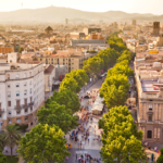 Aerial view of La Rambla, a bustling tree-lined promenade in Barcelona. The scene shows a vibrant mix of pedestrians walking along the street, surrounded by lush green trees. In the background, the cityscape is dotted with buildings, and distant hills provide a scenic backdrop. The atmosphere is lively, capturing the essence of urban life in Barcelona.